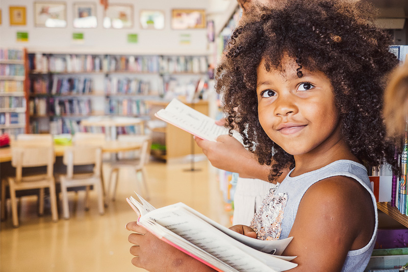 Elementary school children in a library leaning against a wall of books and reading.