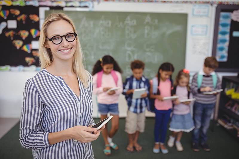 Students lined up and working on devices as teacher smiles with tablet in front of them.
